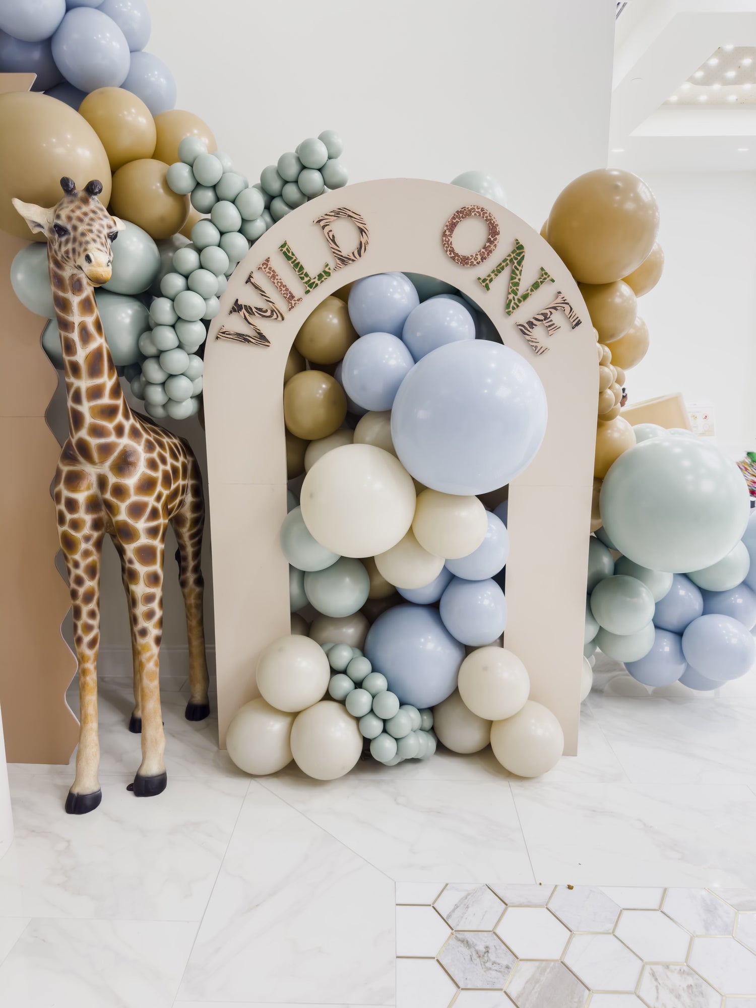 Luxury 'Wild-One' safari themed first birthday party balloon arch with life sized giraffe prop and pastel-colored balloon display in Dallas event venue